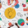 Bir ekip, üzerinde ampul ve dişli simgeleri olan büyük bir diyagram etrafında toplanarak Design Thinking (tasarım odaklı düşünme) sürecinin adımlarını iş birliği içinde planlıyor ve çizim yapıyor.